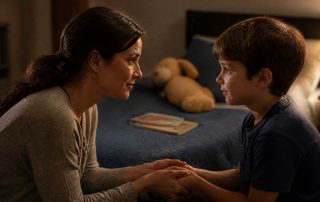 A mother kneels at eye level with her young son, holding his hands as she talks gently in his bedroom