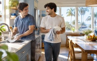 Two young men wash and dry dishes together in a bright, tidy shared kitchen with palm trees outside